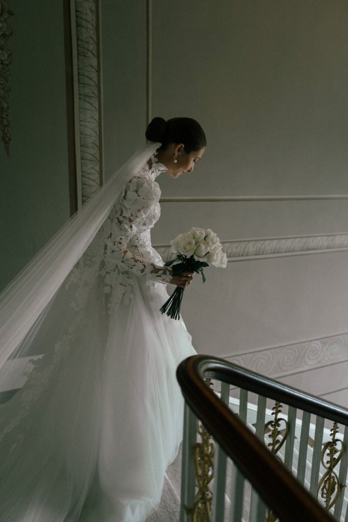 A bride, holding her bouquet walks down the stairs of an English stately home to her ceremony.