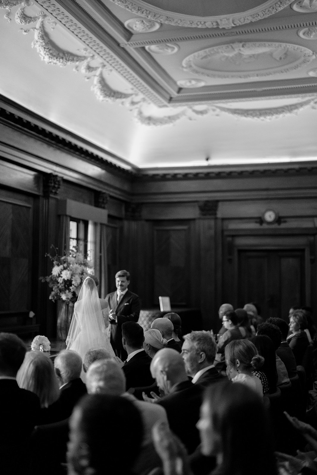 A couple stand holding hands, mid ceremony, at the front of the main room at Old Marylebone Town Hall surrounded by their friends and family.