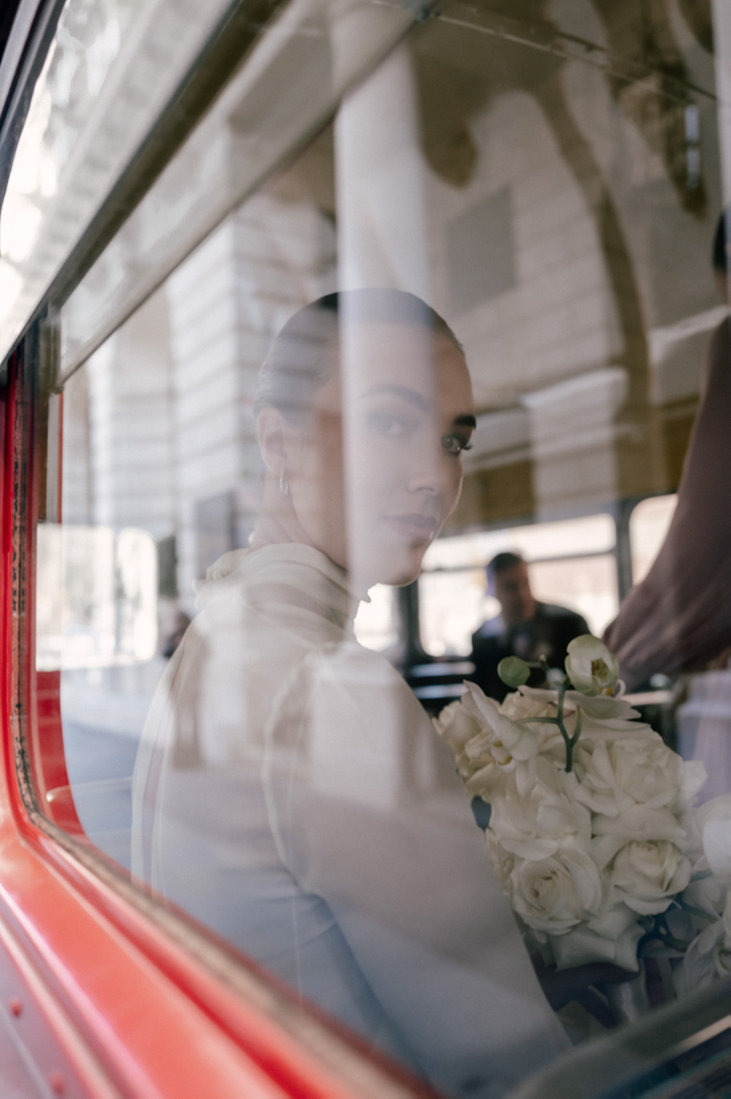 A bride can be seen through the window of a double decker red London bus. She's holding a white bouquet and is looking straight into the camera lens.