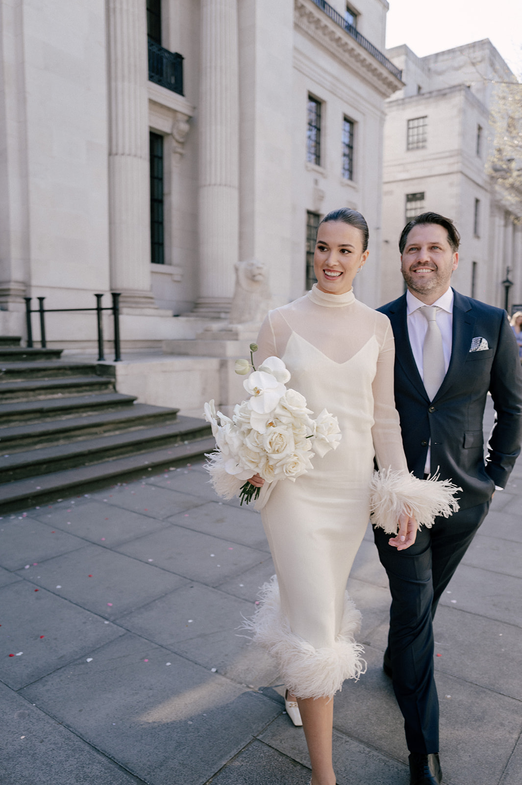 A style bride and groom walk on the pavement outside Old Marylebone Town Hall. The bride, wearing a white dress with long sleeves is holding large white bouquet.