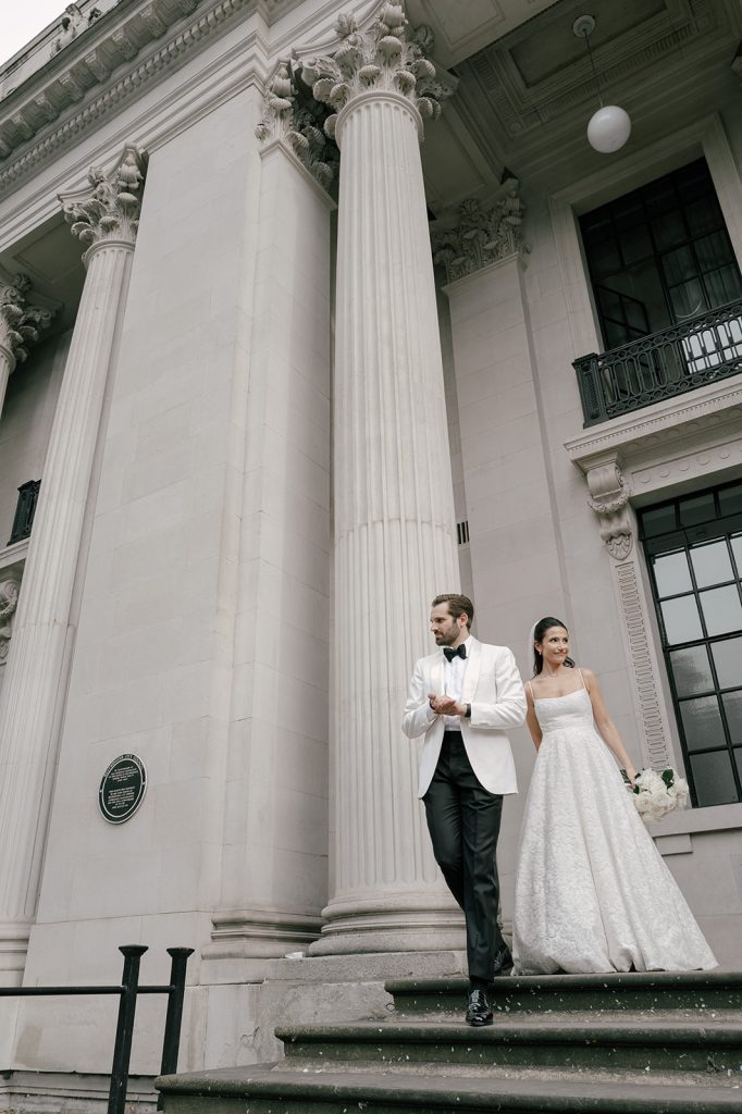 A newly married couple leave Old Marylebone Town Hall following their Wedding Ceremony. The  groom wearing a white jacket and bow tie leads his wife down the stone steps outside of the venue.