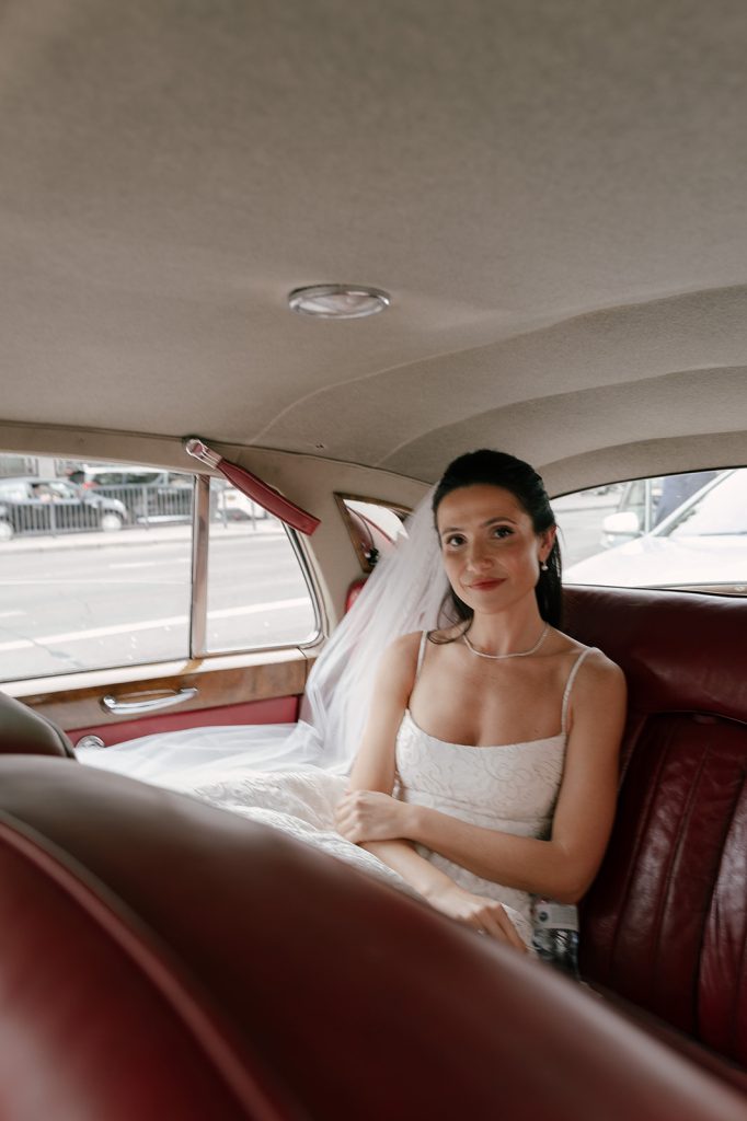 A bride with dark hair and a long veil sits in a car outside of Old Marylebone town hall. 